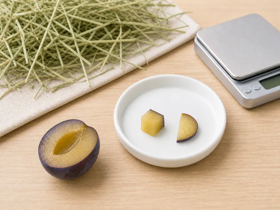 Tiny plum cube and slice on a saucer beside a pitted plum half, hay, water, and a gram scale.